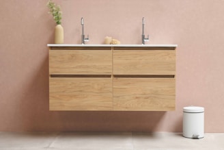 A modern bathroom setting featuring a wooden floating vanity with two chrome faucets. On the left, a tall, beige vase contains green foliage, while a small decorative object sits between the sinks. The background wall is painted in a light, warm tone, and a white, round pedal bin is placed on the floor to the right.