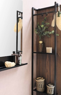 The image depicts a modern bathroom shelf with a minimalist design. There are two bottles of liquid soap with black caps, a natural sponge, and a woven basket placed on the shelf. Above, there is a mirror reflecting part of the room. Next to the shelf is a plant in a glass jar, adding a touch of greenery. The background consists of a wood panel and a light pink wall.