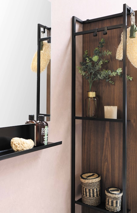 The image depicts a modern bathroom shelf with a minimalist design. There are two bottles of liquid soap with black caps, a natural sponge, and a woven basket placed on the shelf. Above, there is a mirror reflecting part of the room. Next to the shelf is a plant in a glass jar, adding a touch of greenery. The background consists of a wood panel and a light pink wall.