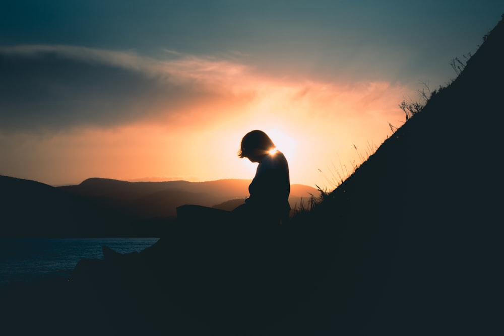 silhouette of man sitting on rock near body of water during sunset