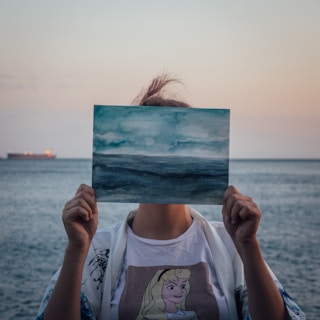 A person holds a painting that depicts a blue and gray seascape, obscuring their face. The background consists of an actual seascape with similar colors, and a ship is visible on the horizon. The person's attire includes a t-shirt with a cartoon character and a light jacket.