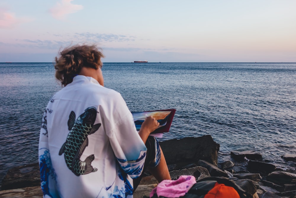 woman in white and black long sleeve shirt sitting on pink textile near body of water