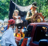 A vibrant street parade features a person dressed as a pirate, complete with an eyepatch, steering a wooden ship's wheel in the bed of a red pickup truck. Nearby, another person in a pirate-themed outfit holds a soda can and a sword, standing confidently. The scene is lively, with people in the background enjoying the festivities under the shade of lush green trees.