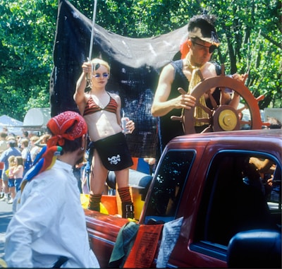 A vibrant street parade features a person dressed as a pirate, complete with an eyepatch, steering a wooden ship's wheel in the bed of a red pickup truck. Nearby, another person in a pirate-themed outfit holds a soda can and a sword, standing confidently. The scene is lively, with people in the background enjoying the festivities under the shade of lush green trees.