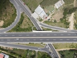 An aerial view of a highway interchange shows multiple roads and lanes with cars moving in different directions. Green patches of vegetation and some buildings are visible around the roads. The main highway runs horizontally across the center of the image, with several connecting roads forming intersections around it.