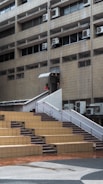 A multi-story building with a facade of beige tiles and numerous air conditioning units attached below the windows. A staircase with white railings leads up from an open courtyard area with stepped seating made of brown and beige tiles. A person is visible near the staircase entrance.
