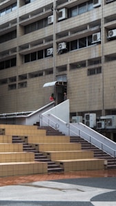 A multi-story building with a facade of beige tiles and numerous air conditioning units attached below the windows. A staircase with white railings leads up from an open courtyard area with stepped seating made of brown and beige tiles. A person is visible near the staircase entrance.