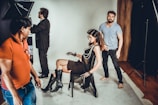 A fashion photoshoot is taking place in a studio. A female model is seated on a chair wearing a sheer black outfit paired with knee-high black boots and layered necklaces. She strikes a pose as several crew members assist with the setup. One person in a red shirt stands in the foreground while another in a blue shirt stands behind the model, adjusting her posture.