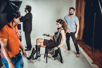 A fashion photoshoot is taking place in a studio. A female model is seated on a chair wearing a sheer black outfit paired with knee-high black boots and layered necklaces. She strikes a pose as several crew members assist with the setup. One person in a red shirt stands in the foreground while another in a blue shirt stands behind the model, adjusting her posture.
