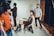 A fashion photoshoot is taking place in a studio. A female model is seated on a chair wearing a sheer black outfit paired with knee-high black boots and layered necklaces. She strikes a pose as several crew members assist with the setup. One person in a red shirt stands in the foreground while another in a blue shirt stands behind the model, adjusting her posture.