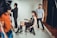 A fashion photoshoot is taking place in a studio. A female model is seated on a chair wearing a sheer black outfit paired with knee-high black boots and layered necklaces. She strikes a pose as several crew members assist with the setup. One person in a red shirt stands in the foreground while another in a blue shirt stands behind the model, adjusting her posture.
