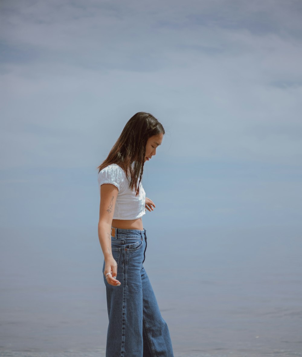 woman in white shirt and blue denim jeans standing on seashore during daytime