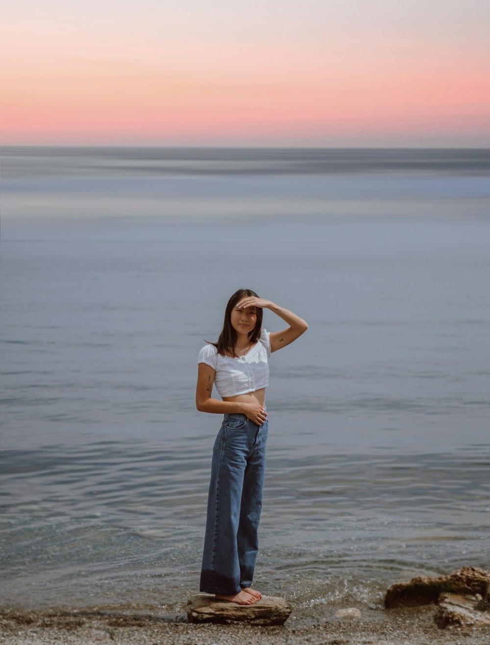 woman in white tank top and blue denim jeans standing on sea shore during daytime