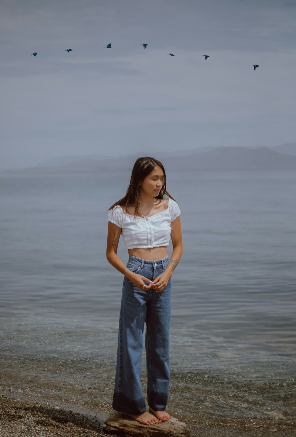woman in white shirt and blue denim jeans standing on beach during daytime