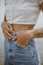 A person wearing a white textured crop top and denim high-waisted jeans. The person has tattoos on their arm and is adjusting the waistband with their hand, which has rings and light blue nail polish.