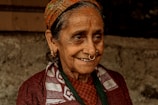 An elderly woman with a warm smile, wearing a traditional patterned headscarf, hoop earrings, and a nose ring. Her clothing includes a patterned scarf over a dark sweater. Her face shows signs of age, with wrinkles and a gentle expression, set against a blurred, earthy background.