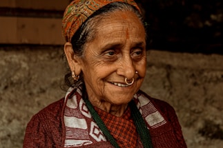 An elderly woman with a warm smile, wearing a traditional patterned headscarf, hoop earrings, and a nose ring. Her clothing includes a patterned scarf over a dark sweater. Her face shows signs of age, with wrinkles and a gentle expression, set against a blurred, earthy background.