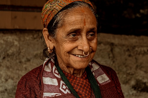 An elderly woman with a warm smile, wearing a traditional patterned headscarf, hoop earrings, and a nose ring. Her clothing includes a patterned scarf over a dark sweater. Her face shows signs of age, with wrinkles and a gentle expression, set against a blurred, earthy background.