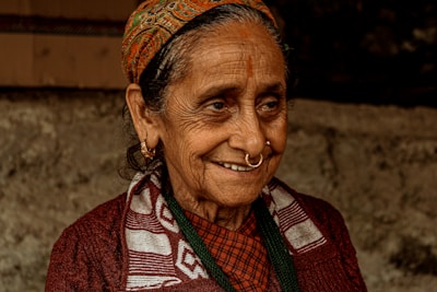An elderly woman with a warm smile, wearing a traditional patterned headscarf, hoop earrings, and a nose ring. Her clothing includes a patterned scarf over a dark sweater. Her face shows signs of age, with wrinkles and a gentle expression, set against a blurred, earthy background.