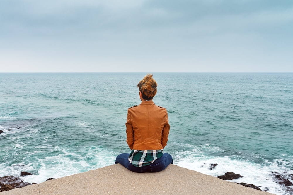 Donna in giacca marrone che si siede sulla roccia marrone vicino al mare durante il giorno