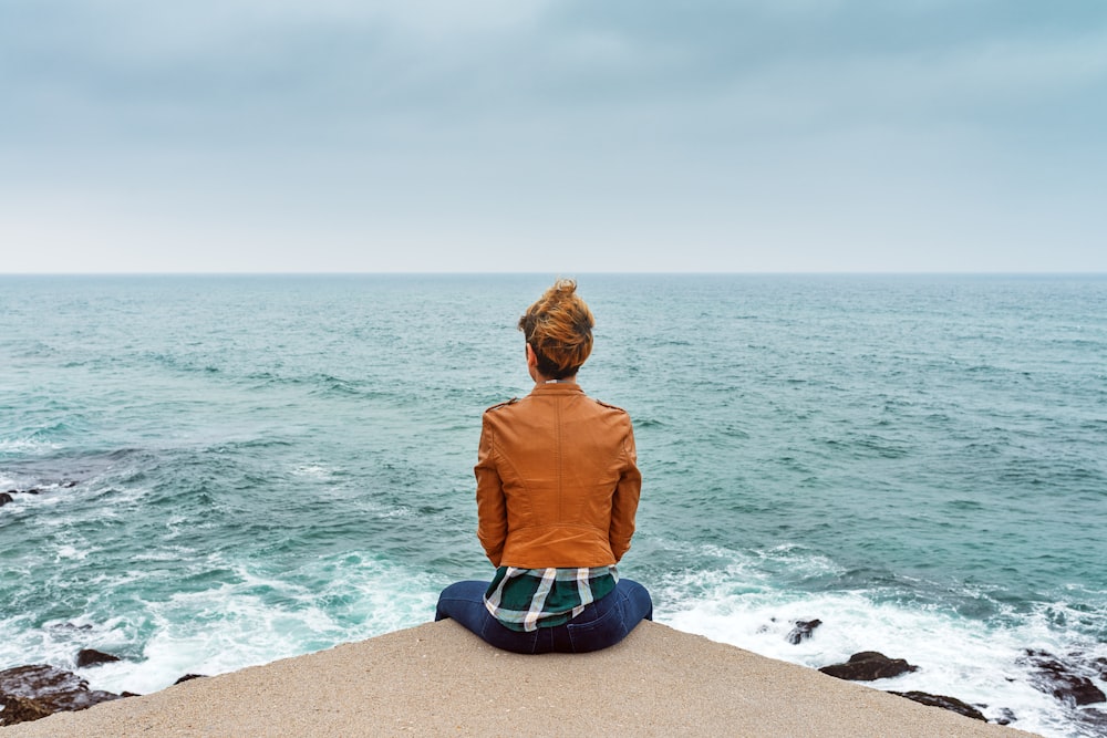 woman in brown jacket sitting on brown rock near sea during daytime