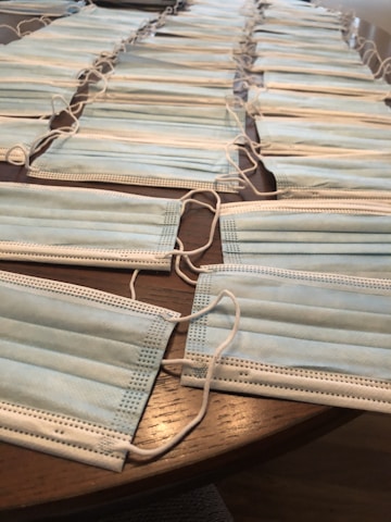 Multiple blue disposable face masks are neatly arranged in rows on a wooden table. The masks have white ear loops attached to them.