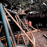A playground scene featuring metal play structures with slides and swings. A person and a child are interacting near one of the structures, which is surrounded by lush green foliage.