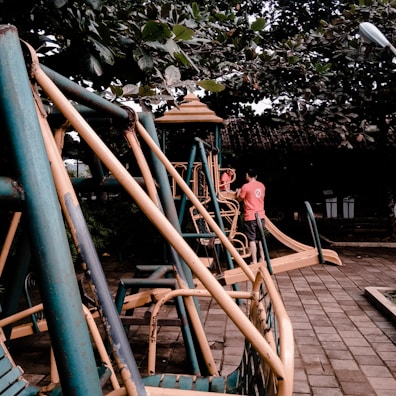 A playground scene featuring metal play structures with slides and swings. A person and a child are interacting near one of the structures, which is surrounded by lush green foliage.