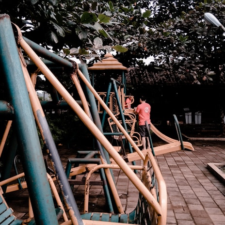 A playground scene featuring metal play structures with slides and swings. A person and a child are interacting near one of the structures, which is surrounded by lush green foliage.