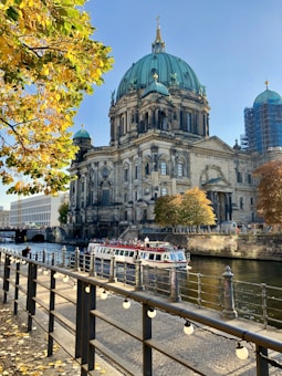 A grand historic building with a large green dome stands prominently by a river. The scene is framed by autumnal trees with yellow leaves, and a sightseeing boat populated with tourists passes along the water. A black railing adorned with small white lights borders the cobblestone walkway beside the river.