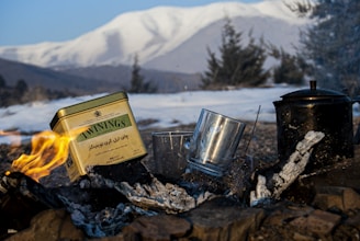 A close-up view of a campfire with a tin of Twinings tea prominently placed. There are two transparent glass cups and a blackened kettle next to the fire. In the background, snow-covered mountains and coniferous trees create a serene outdoor setting.