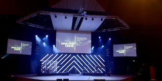 A stage setup with a large screen displaying 'JOIN THE CREW.' The stage is equipped with lighting fixtures, speakers, and musical instruments. There are two side screens with the same message. Blue and white lights create a vibrant atmosphere.