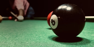 A close-up view of an eight-ball on a green pool table, with a cue stick and several other balls in the background. The focus is on the black eight-ball, with a red ball just behind it. A person is positioned in the background ready to take a shot.