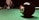 A close-up view of an eight-ball on a green pool table, with a cue stick and several other balls in the background. The focus is on the black eight-ball, with a red ball just behind it. A person is positioned in the background ready to take a shot.