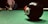 A close-up view of an eight-ball on a green pool table, with a cue stick and several other balls in the background. The focus is on the black eight-ball, with a red ball just behind it. A person is positioned in the background ready to take a shot.