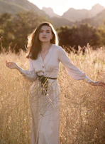 A model wearing a flowing western dress with delicate embroidery standing in a sunlit field.