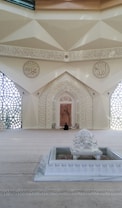 A beautifully decorated interior space blends classical Islamic architectural elements with modern design. Intricate geometric patterns adorn large windows, allowing light to filter through, casting intricate shadows on the floor. Ornate Arabic calligraphy decorates the walls, adding to the spiritual and cultural ambiance of the space. A small fountain or decorative water feature sits prominently in the center, enhancing the serene atmosphere.