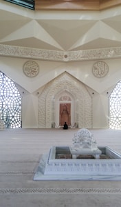 A beautifully decorated interior space blends classical Islamic architectural elements with modern design. Intricate geometric patterns adorn large windows, allowing light to filter through, casting intricate shadows on the floor. Ornate Arabic calligraphy decorates the walls, adding to the spiritual and cultural ambiance of the space. A small fountain or decorative water feature sits prominently in the center, enhancing the serene atmosphere.