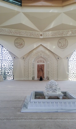 A beautifully decorated interior space blends classical Islamic architectural elements with modern design. Intricate geometric patterns adorn large windows, allowing light to filter through, casting intricate shadows on the floor. Ornate Arabic calligraphy decorates the walls, adding to the spiritual and cultural ambiance of the space. A small fountain or decorative water feature sits prominently in the center, enhancing the serene atmosphere.