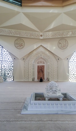 A beautifully decorated interior space blends classical Islamic architectural elements with modern design. Intricate geometric patterns adorn large windows, allowing light to filter through, casting intricate shadows on the floor. Ornate Arabic calligraphy decorates the walls, adding to the spiritual and cultural ambiance of the space. A small fountain or decorative water feature sits prominently in the center, enhancing the serene atmosphere.