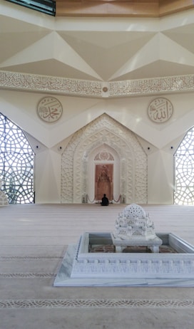 A beautifully decorated interior space blends classical Islamic architectural elements with modern design. Intricate geometric patterns adorn large windows, allowing light to filter through, casting intricate shadows on the floor. Ornate Arabic calligraphy decorates the walls, adding to the spiritual and cultural ambiance of the space. A small fountain or decorative water feature sits prominently in the center, enhancing the serene atmosphere.