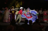 A group of people dressed in traditional, colorful attire is performing a dance on stage. The central figure is a woman wearing a bright blue and red dress with a wide, flowing skirt, while a man in a red shirt and beige pants dances beside her. Other performers in the background wear a variety of traditional costumes, including sombreros and dresses with floral designs.