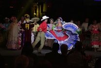 A group of people dressed in traditional, colorful attire is performing a dance on stage. The central figure is a woman wearing a bright blue and red dress with a wide, flowing skirt, while a man in a red shirt and beige pants dances beside her. Other performers in the background wear a variety of traditional costumes, including sombreros and dresses with floral designs.