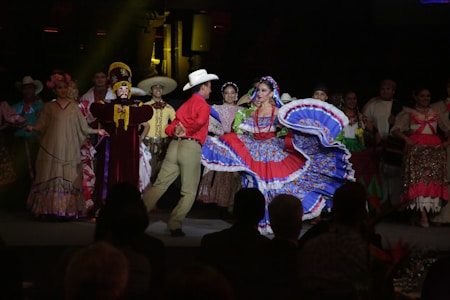 A group of people dressed in traditional, colorful attire is performing a dance on stage. The central figure is a woman wearing a bright blue and red dress with a wide, flowing skirt, while a man in a red shirt and beige pants dances beside her. Other performers in the background wear a variety of traditional costumes, including sombreros and dresses with floral designs.