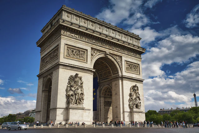 The Arc de Triomphe seen at an angle against a partly cloudy sky with traffic circling below
