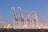 Four large industrial cranes stand tall on a dockside, silhouetted against a twilight sky. The background features city lights and buildings, creating a contrast with the industrial scene in the foreground.