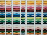 A neatly organized assortment of colorful paper sheets is displayed on shelves. Each row contains papers of a different color, forming a gradient from light to dark shades. The shelves are structured in columns, creating a visually appealing and orderly arrangement.