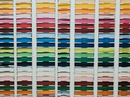 A neatly organized assortment of colorful paper sheets is displayed on shelves. Each row contains papers of a different color, forming a gradient from light to dark shades. The shelves are structured in columns, creating a visually appealing and orderly arrangement.