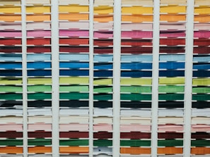 A neatly organized assortment of colorful paper sheets is displayed on shelves. Each row contains papers of a different color, forming a gradient from light to dark shades. The shelves are structured in columns, creating a visually appealing and orderly arrangement.