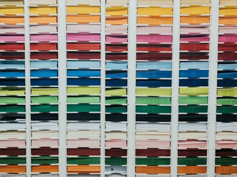 A neatly organized assortment of colorful paper sheets is displayed on shelves. Each row contains papers of a different color, forming a gradient from light to dark shades. The shelves are structured in columns, creating a visually appealing and orderly arrangement.